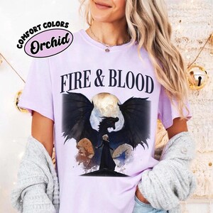 May include: A light purple short-sleeve t-shirt featuring the text "FIRE & BLOOD" in black letters above a graphic of a dark dragon with large wings and a figure in a black cloak standing before a full moon, with a "COMFORT COLORS Orchid" label.