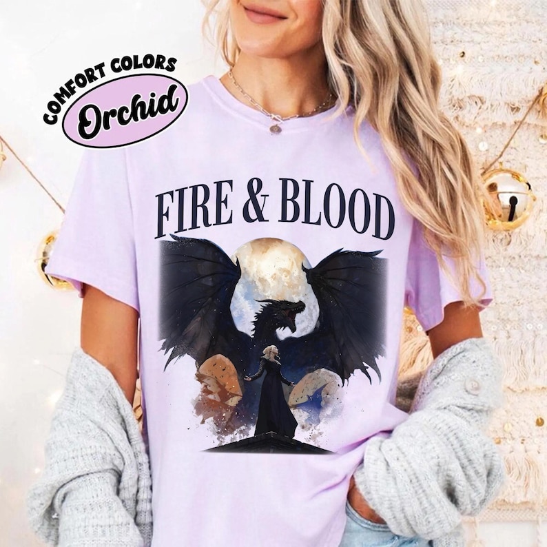May include: A light purple short-sleeve t-shirt featuring the text "FIRE & BLOOD" in black letters above a graphic of a dark dragon with large wings and a figure in a black cloak standing before a full moon, with a "COMFORT COLORS Orchid" label.