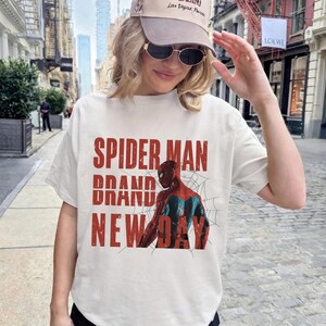 K&ouml;nnte beinhalten: Wei&szlig;es T-Shirt mit einer Spider-Man-Grafik und den Worten "SPIDER MAN BRAND NEW DAY" in Rot. Das Shirt wird mit einer beigen Kappe und Sonnenbrille getragen. Das Thema ist Comic und Superheld.