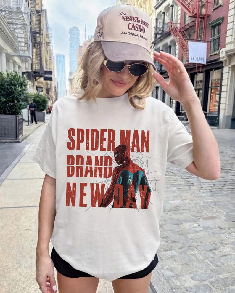 K&ouml;nnte beinhalten: Wei&szlig;es T-Shirt mit einer Spider-Man-Grafik und den Worten "SPIDER MAN BRAND NEW DAY" in Rot. Das Shirt wird mit einer beigen Kappe und Sonnenbrille getragen. Das Thema ist Comic und Superheld.