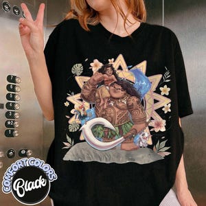 May include: A black t-shirt featuring a graphic print of Moana and Maui from the movie Moana. Maui is holding a large white hook, with a small rooster and pig nearby. The design includes a yellow star-shaped background with tropical flowers.