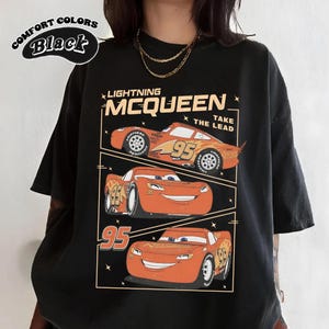 May include: Black t-shirt featuring Lightning McQueen from the Cars movie. The design includes three images of the character with the number 95 and the text "Lightning McQueen Take The Lead". The shirt also has the text "Comfort Colors Black".