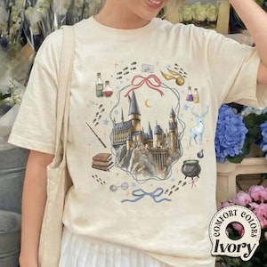 May include: An ivory t-shirt featuring a central graphic of a castle surrounded by a pink ribbon, a golden snitch, a stag, a cauldron, books, a wand, and potion bottles. The design includes scattered footprints and a blue ribbon at the bottom.