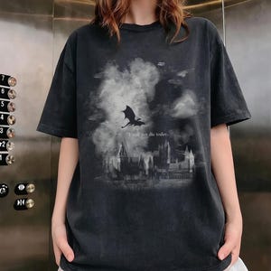 May include: Dark grey t-shirt with a dragon silhouette flying over a castle scene and cloudy sky. The text "I will not die today" is printed below the dragon. The shirt has short sleeves and a crew neck.