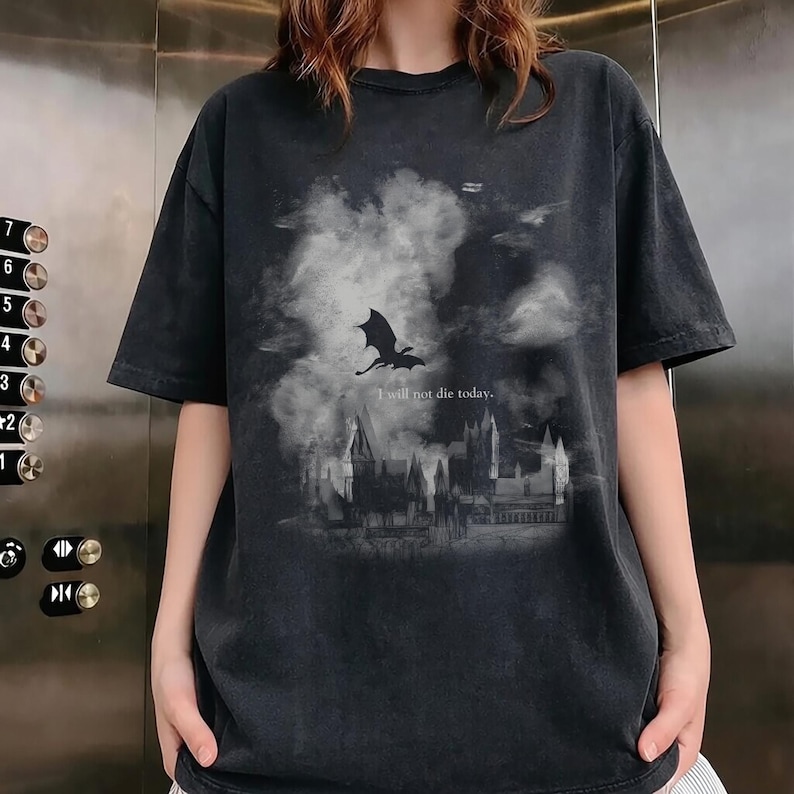 May include: Dark grey t-shirt with a dragon silhouette flying over a castle scene and cloudy sky. The text "I will not die today" is printed below the dragon. The shirt has short sleeves and a crew neck.
