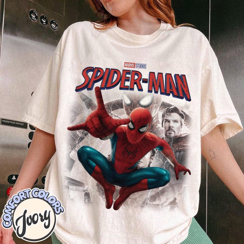 May include: An ivory short-sleeve t-shirt featuring a graphic print of Spider-Man in a dynamic pose, with the text "MARVEL STUDIOS SPIDER-MAN" above him. The design includes a background image of Spider-Man's mask and a portrait of Doctor Strange.
