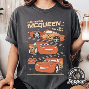 May include: Dark gray t-shirt featuring a graphic of Lightning McQueen from the Cars movie. The design includes three images of the red race car with the number 95, along with the text "Lightning McQueen Take The Lead."