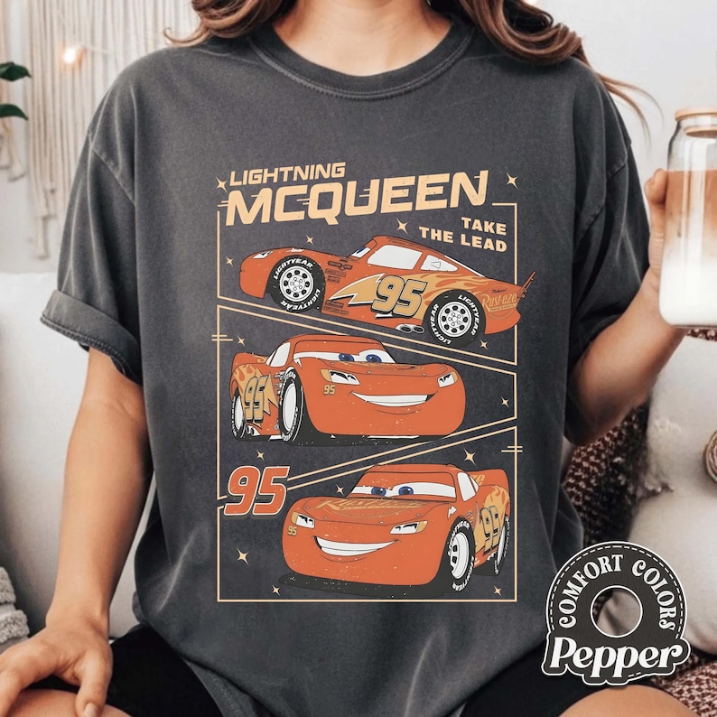 May include: Dark gray t-shirt featuring a graphic of Lightning McQueen from the Cars movie. The design includes three images of the red race car with the number 95, along with the text "Lightning McQueen Take The Lead."