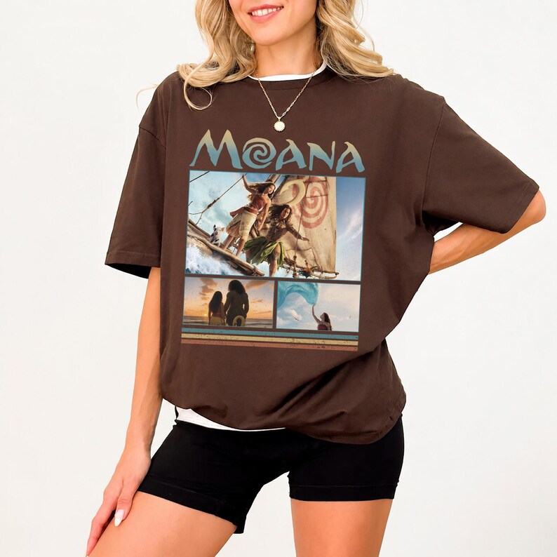 May include: A dark brown short-sleeve t-shirt featuring a graphic print with the text MOANA at the top. The design includes three separate scenes from the movie Moana, showing characters on a boat, standing on a beach, and holding a blue cloth.