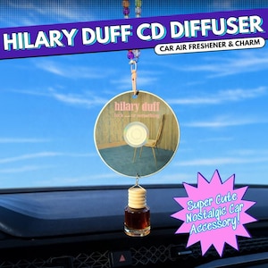 May include: A car air freshener and charm featuring a CD design with "HILARY DUFF" printed on it. The CD has an image of a room with a chair. A small bottle of liquid hangs below the CD.