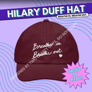 May include: A maroon baseball cap featuring the white embroidered text "Breathe in. Breathe out." with a small white heart icon. The cap has a curved brim and a structured crown with ventilation eyelets, presented against a purple background.