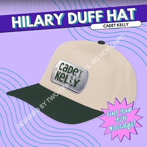 May include: A beige baseball cap with a dark green brim. The front of the cap features a silver-colored rectangular metal tag with the text "CADET KELLY" printed in dark green letters. The cap has a dark green button at the top center.