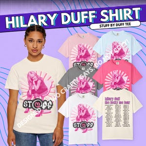 May include: A collection of short-sleeve t-shirts in various colors including cream, light blue, pink, and gray. Each shirt features a graphic of a person sitting on a chair with the text STUFF and the Hilary Duff The Lucky Me Tour list.