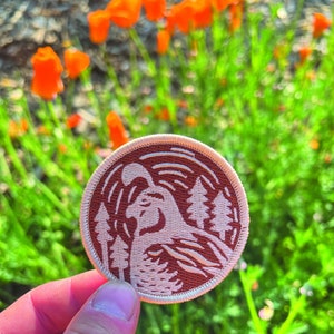 Puede incluir: Un parche bordado marrón y blanco con una imagen estilizada de un pájaro en un entorno de bosque. El parche está rodeado por un borde blanco.