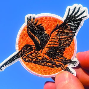 May include: Embroidered patch featuring a pelican in flight, with black and orange details. The pelican is set against an orange circle, with a white border. The patch is held against a blue sky background.