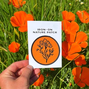 May include: An iron-on nature patch featuring a black floral design on an orange circle. The patch is held in front of a background of bright orange flowers and green grass. The text "IRON-ON NATURE PATCH" is printed above the patch.