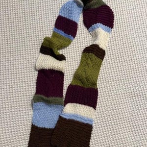 Op de afbeelding: Een handgebreide sjaal met een gestreept patroon. De sjaal heeft secties in bruin, blauw, wit, groen en bordeaux. De sjaal is gemaakt van een zacht, getextureerd garen en is ontworpen om om de nek te worden gedragen.