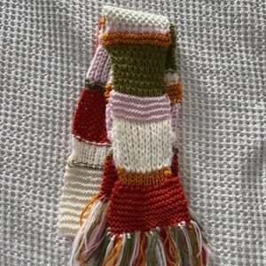 Op de afbeelding: Een handgebreide sjaal met een kleurrijk streepjespatroon. De sjaal heeft rode, witte, groene, roze en oranje strepen. De uiteinden zijn afgewerkt met franjes in bijpassende kleuren. Een gezellig winteraccessoire.