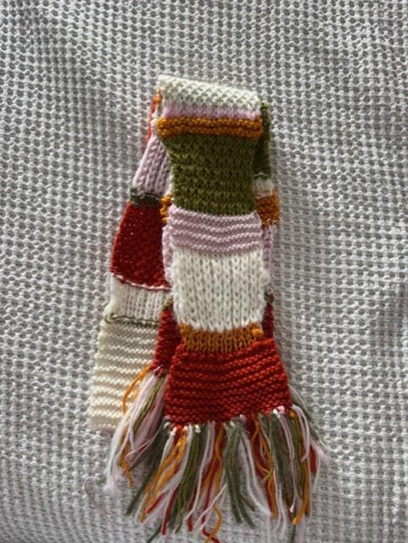 Op de afbeelding: Een handgebreide sjaal met een kleurrijk streepjespatroon. De sjaal heeft rode, witte, groene, roze en oranje strepen. De uiteinden zijn afgewerkt met franjes in bijpassende kleuren. Een gezellig winteraccessoire.