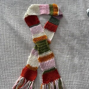 Op de afbeelding: Een kleurrijke gebreide sjaal met een gestreept patroon. De sjaal heeft verschillende kleuren, waaronder rood, wit, roze, oranje, groen en paars. De uiteinden van de sjaal hebben franjes. De sjaal is gemaakt van zacht, getextureerd garen.