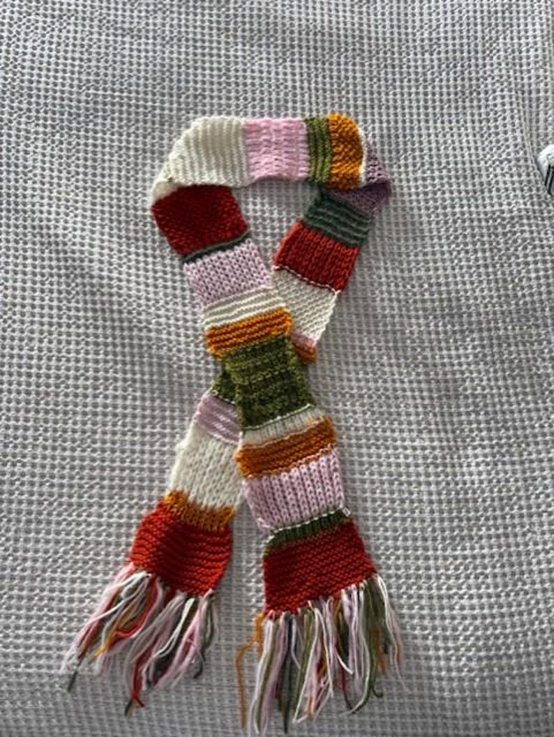 Op de afbeelding: Een kleurrijke gebreide sjaal met een gestreept patroon. De sjaal heeft verschillende kleuren, waaronder rood, wit, roze, oranje, groen en paars. De uiteinden van de sjaal hebben franjes. De sjaal is gemaakt van zacht, getextureerd garen.