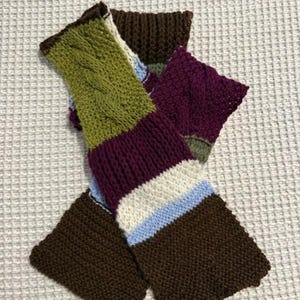 Op de afbeelding: Een kleurrijke, handgebreide sjaal met verschillende kleuren. De sjaal heeft secties in bruin, groen, paars, wit en blauw. Het groene gedeelte heeft een kabelbreipatroon. De sjaal is gemaakt van zacht, getextureerd garen.