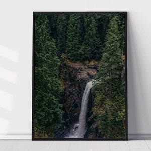 May include: A framed print depicting a waterfall plunging into a gorge, flanked by dense, green coniferous trees. The artwork is presented in a black frame, offering a striking contrast to the natural landscape.