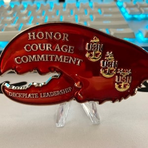 May include: A red, lobster claw-shaped challenge coin with the words "HONOR COURAGE COMMITMENT" and "DECKPLATE LEADERSHIP" in silver lettering. The coin features three gold USN anchor emblems.