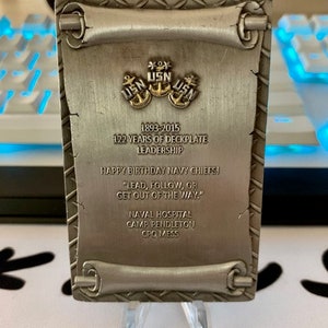 Puede incluir: Placa rectangular plateada con el texto "1893-2015 122 YEARS OF DECKPLATE LEADERSHIP" y "HAPPY BIRTHDAY NAVY CHIEFS!". La placa presenta tres anclas USN doradas y diseños en forma de pergamino en la parte superior e inferior.