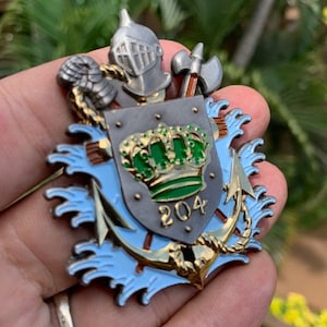 Puede incluir: Un emblema metálico detallado con un yelmo de caballero, un hacha y un escudo con una corona y el número 204. El emblema tiene detalles dorados, plateados, verdes y azules. El emblema se sostiene en una mano.