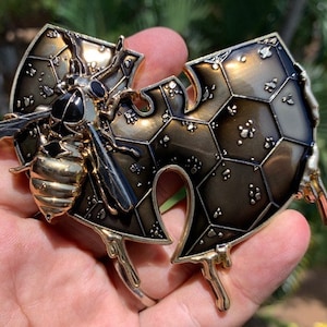 May include: A detailed, bronze-toned metal bracelet featuring a bee and honeycomb design. The bee has black and gold accents, and the honeycomb is made of geometric shapes. The bracelet has a unique, artistic style.