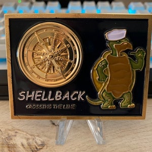 May include: A rectangular, gold-framed coin with a black background. The coin features a gold compass rose and a cartoon turtle wearing a sailor hat. The text "SHELLBACK CROSSING THE LINE" is displayed in silver lettering.