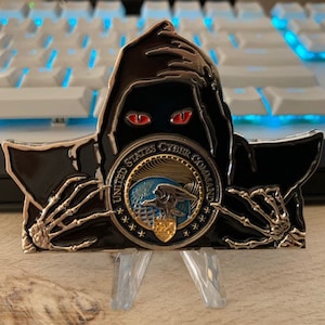 May include: A black and gold United States Cyber Command coin featuring a grim reaper design with red eyes and skeletal hands. The coin has the text "UNITED STATES CYBER COMMAND" and a globe and eagle emblem.