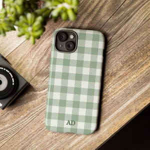 May include: A sage green and white gingham pattern phone case with the initials "AD" at the bottom. The phone case is on a wooden surface next to a camera and greenery.