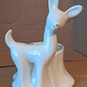 May include: A white ceramic deer planter with a hollowed-out back. The deer has a slender neck, upright ears, and a curved tail. The planter is in the shape of a tree stump, and the deer is standing in front of it.