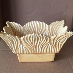 May include: A decorative, square-shaped bowl with a floral design. The bowl is made of a light tan material and features a petal-like rim with vertical grooves. The base is a solid, rectangular shape. The bowl is empty.