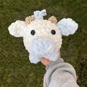 May include: A crocheted stuffed cow toy with white fur, blue ears, and brown horns. The cow has black eyes and a light blue nose.