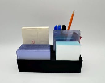 May include: A black desk organizer with compartments holding a stack of light purple cards, a white block, a clear container with pens and a pencil, and a manila envelope. The organizer is on a white surface.