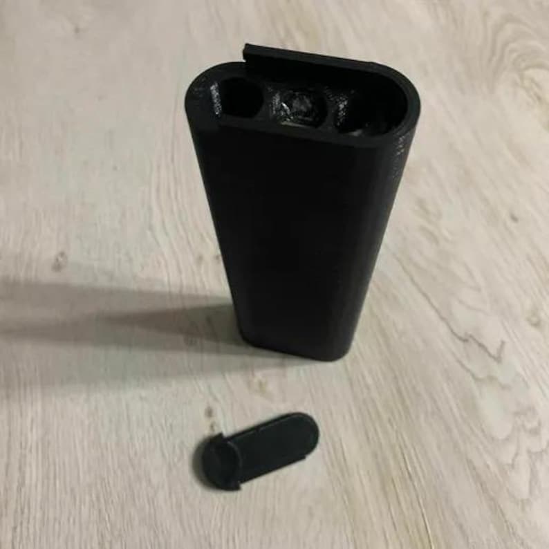 Op de afbeelding: Een zwarte, taps toelopende container met drie cirkelvormige openingen aan de bovenkant en een kleine, losgemaakte, zwarte dop. De container is gemaakt van een mat materiaal en staat op een lichtgekleurd houten oppervlak.