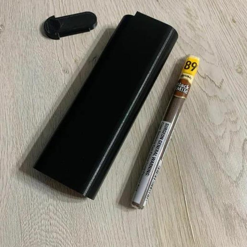 Op de afbeelding: Een zwarte rechthoekige koffer met een apart deksel en een sigaar. De sigaar is gemarkeerd met "Black Mild" en heeft een gele bovenkant met het nummer "89". De koffer en het deksel zijn gemaakt van matzwart materiaal.