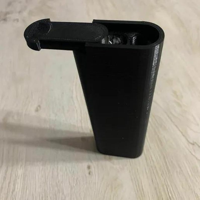 Op de afbeelding: Een zwarte, rechthoekige container met scharnieren, een gebogen bovenkant en een deksel dat opengaat. De container is gemaakt van een solide, mat materiaal en staat op een lichtgekleurd houten oppervlak. Het deksel is open en onthult de binnenkant.