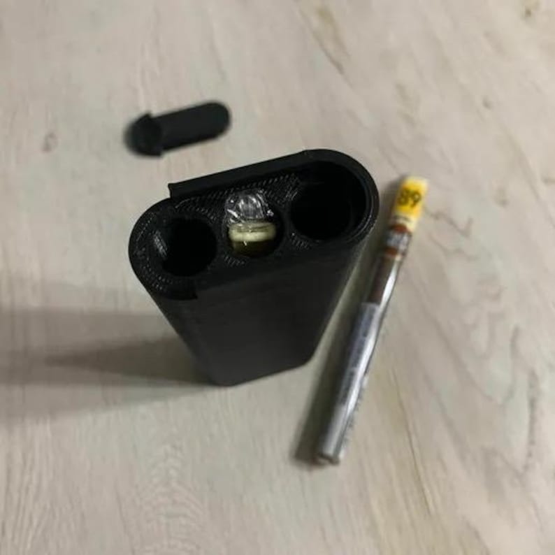 Op de afbeelding: Zwarte rechthoekige koffer met drie openingen, waarvan &eacute;&eacute;n met een klein lampje. Een zwarte dop is in de buurt. Er is ook een sigaar met een geel en zilveren label.