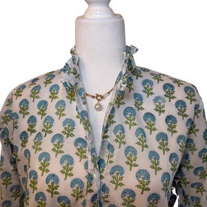 May include: A white button-down shirt with a blue and green floral print. The shirt has a ruffled collar and a pearl necklace.