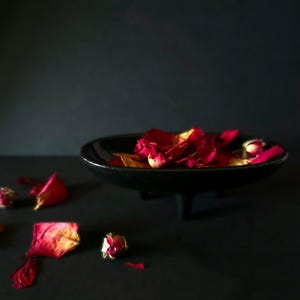 May include: Dried rose petals in a black bowl with a black background. The petals are a deep red color and some have a yellow tint. There are also a few rose buds scattered around the bowl.