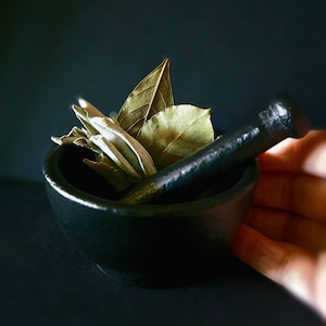 May include: A black mortar and pestle with dried bay leaves and white sage inside.