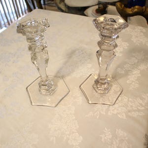 May include: Two clear glass candle holders with hexagonal bases and faceted stems. The candle holders have a classic design and are displayed on a white floral tablecloth. They are suitable for decorative purposes.
