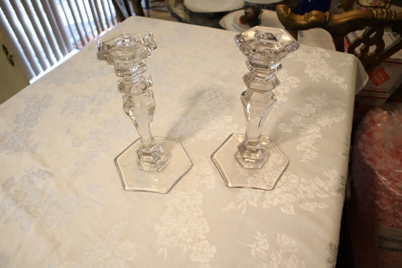 May include: Two clear glass candle holders with hexagonal bases and faceted stems. The candle holders have a classic design and are displayed on a white floral tablecloth. They are suitable for decorative purposes.