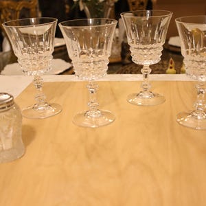 May include: Four clear glass goblets with faceted stems and textured bowls featuring a diamond pattern, accompanied by a small clear glass shaker with a silver-coloured metal lid, all arranged on a flat wooden surface for display.