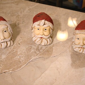 May include: Three Santa Claus head figurines with red hats and white beards are arranged on a marble surface. The Santa heads have detailed facial features and are made of a cream-colored material. The hats have a red finish.