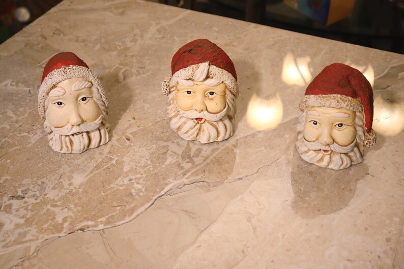 May include: Three Santa Claus head figurines with red hats and white beards are arranged on a marble surface. The Santa heads have detailed facial features and are made of a cream-colored material. The hats have a red finish.